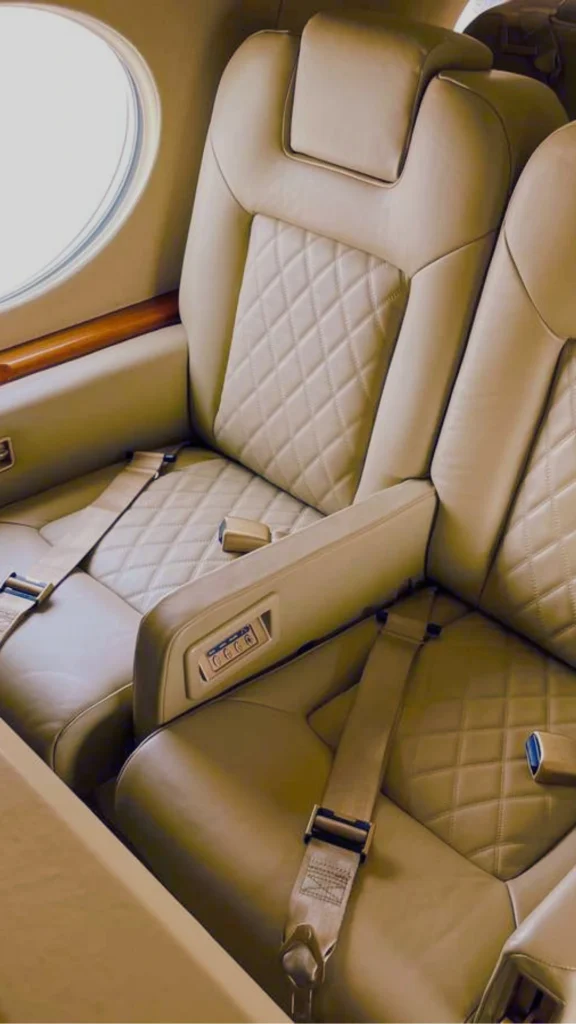 Interior view of luxury private jet seating featuring two beige leather armchairs with intricate diamond stitching on the cushions. The seats are equipped with tan safety seatbelts and electronic control buttons integrated into the center armrest, with a classic airplane window and polished wood trim visible in the background.