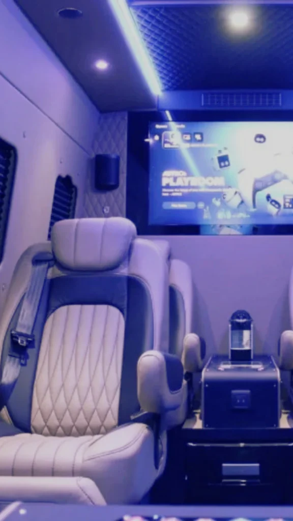 Interior view of a luxury vehicle or private transport cabin designed as a mobile media suite. Features an oversized plush leather captain's chair with white diamond-stitch detailing and a blue safety harness. A sleek media monitor glows in the background, complemented by modern overhead lighting and a compact console table with a beverage or coffee maker.