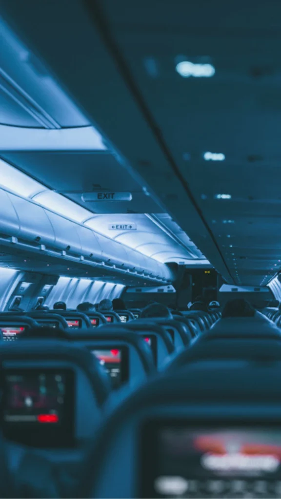 The commercial passenger cabin of an airplane looking down the rows of seats at night, showing the cold, flat blue and white overhead LED lighting that the blog post contrasts against warm, layered luxury aircraft lighting. Passengers sit in rows with illuminated seatback entertainment screens visible in the dimmed environment.
