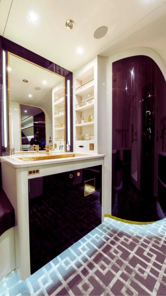 An ultra-luxury private jet spa bathroom showing a residential-style layout. The space features a vanity with a white countertop and a polished gold sink basin, a large gold-trimmed mirror with integrated side lighting, built-in shelving for amenities, and a curved, glossy deep purple shower enclosure. The floor is covered in a geometric-patterned gray carpet lit by subtle under-cabinet glow.
