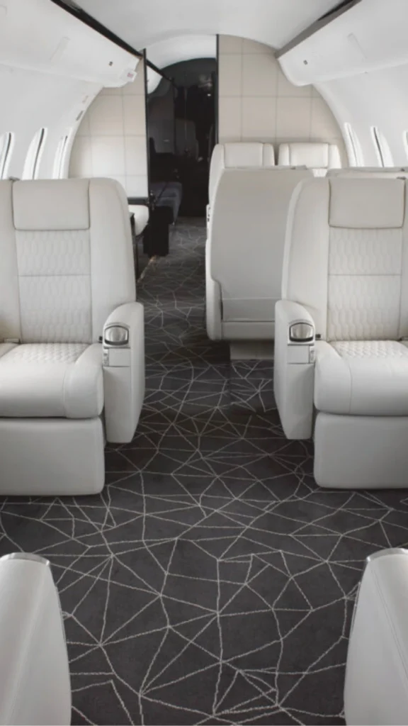 Interior view of a custom-designed private jet cabin showing a dining suite configuration. The space features an elegant, glossy wood table with formal place settings, neutral fabric seating, and a row of illuminated airplane windows. Custom wood trim accents line the curved fuselage, with leather armchairs visible in the background lounge area.