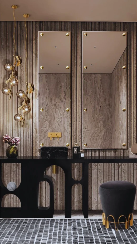 A high-end luxury bathroom vanity area showcasing a rich mix of materials and textures. Two tall rectangular mirrors with prominent brass corner studs are mounted against a textured stone or wood-slat accent wall. A modern black marble basin sits on a long black console table, complemented by an artistic gold branch pendant light, gold fixtures, and a plush dark gray vanity stool with a gold base.
