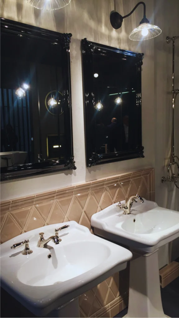 A luxury vintage-style double vanity setup featuring two white ceramic pedestal sinks with polished chrome faucets. Two ornate, glossy black-framed mirrors are mounted on the wall above, separated by a beige diamond-patterned tiled backsplash. A gooseneck wall sconce casts warm lighting over the elegant display.