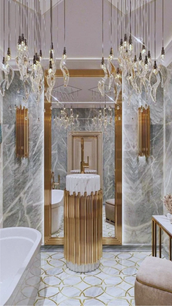 An ultra-luxury bathroom showcasing a highly stylized, layered lighting design. A multitude of delicate, glass pendant lights hang like a sculptural installation from the ceiling, reflecting in a massive, floor-to-ceiling gold-framed mirror. The light illuminates a freestanding white marble pedestal sink encased in vertical gold tubes, modern gold wall sconces, and gray marble walls.