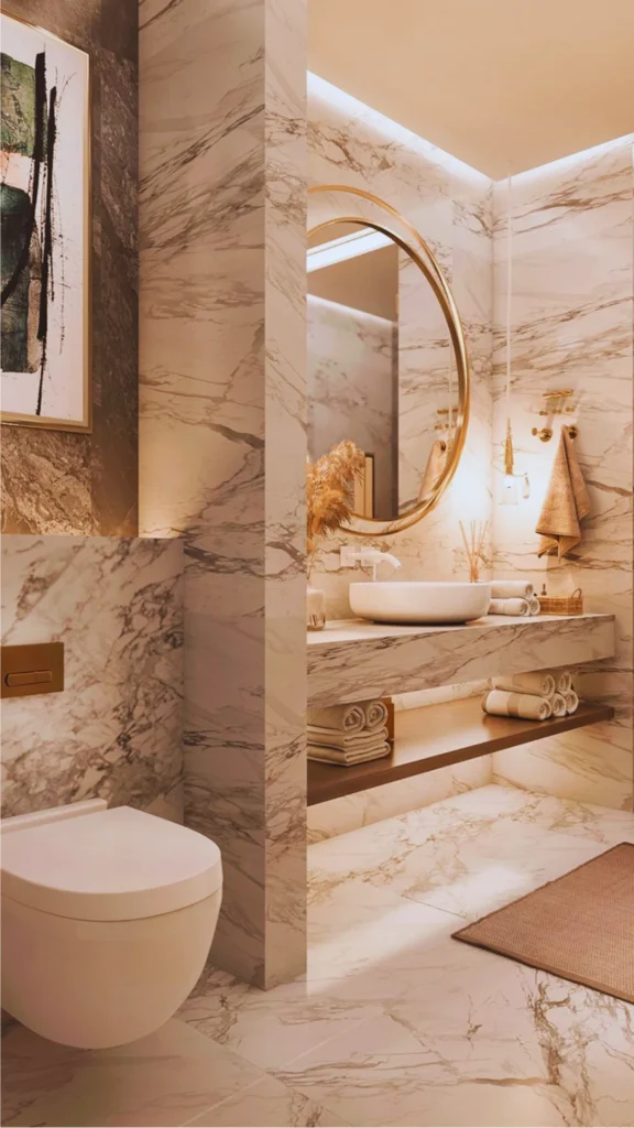Interior view of a high-end modern bathroom showcasing an architectural layout. A marble partition wall partially separates a sleek wall-mounted white toilet from a floating single-sink vanity station. Warm hidden LED strip lighting highlights the detailed marble veins across the walls and floors, while a round brass mirror and hanging pendant add delicate accents.