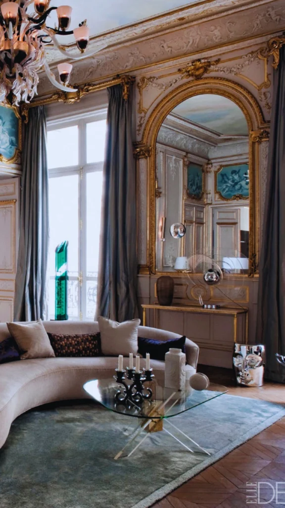 A grand Parisian apartment salon showcasing classic 19th-century Haussmann architectural features mixed with modern furniture. The room boasts ultra-high ceilings with ornate crown molding, a massive gold leaf arched mirror, a tall floor-to-ceiling window framed by long drapes, and traditional herringbone parquet wood flooring. In the foreground, a contemporary curved sofa sits around a modern glass coffee table on a muted blue area rug.