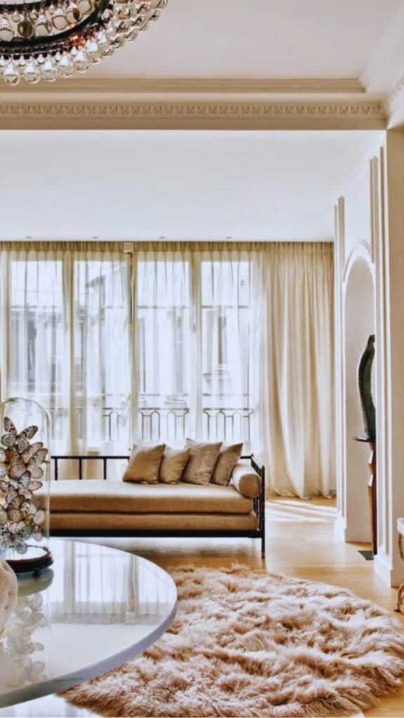 A bright Parisian living room demonstrating the artful blending of eras and textures. A low, vintage-style tan daybed with an intricate dark frame rests in front of a wide floor-to-ceiling window lined with sheer white curtains. The space features an eclectic mix of textures, including a high-pile shaggy round rug, a glossy round white table holding a glass butterfly cloche, and an architectural arched wall nook displaying a contemporary sculpture.