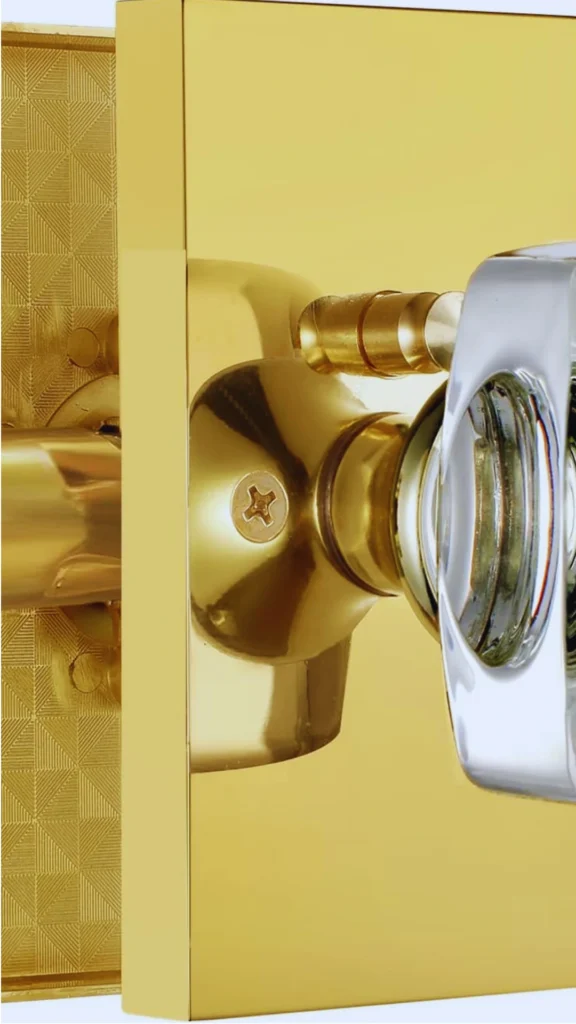 An extreme close-up of a modern premium door knob assembly showcasing minimalist lines and a luxury mix of materials. The architectural fixture features a highly reflective, polished gold square backplate paired with a clear, sculpted glass or crystal knob handle rimmed in polished silver chrome, highlighting the heavy weight and fine detail of the mechanics underneath.