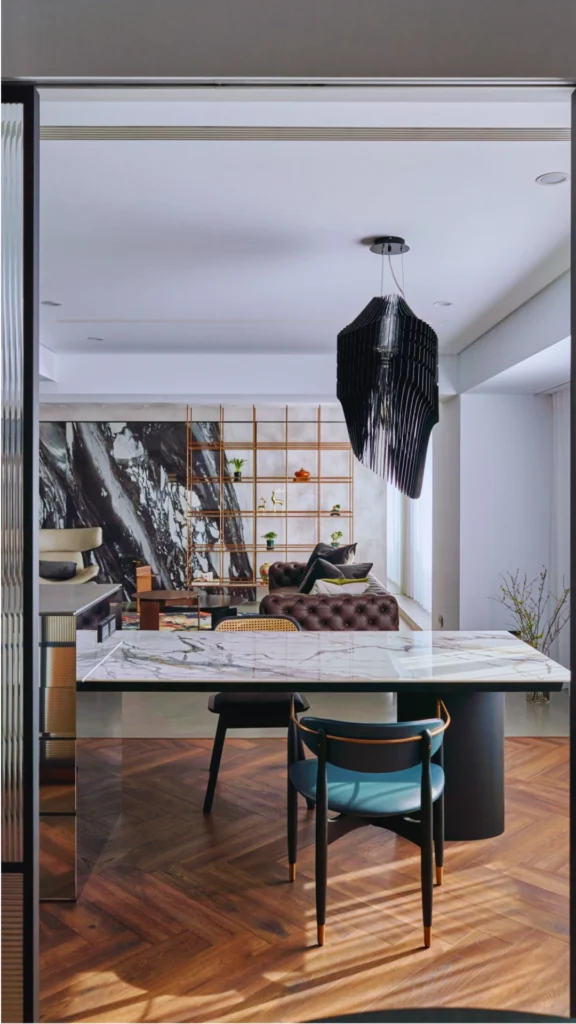 A sleek, open-concept modern home displaying a bold linear and sculptural rhythm. In the foreground, a dining island with a dramatic white-and-gray veined marble slab countertop is paired with contemporary teal and black dining chairs. Hanging above is an oversized, tiered black geometric statement chandelier that anchors the space, framed by rich herringbone wood flooring and a glimpse into a living room with a tufted leather sofa and a marble feature wall.