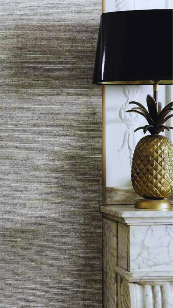 A close-up interior vignette showcasing a beautiful interplay of tactile textures and metallic contrast. A textured, woven grasscloth wallpaper in muted earthy green and gold tones runs alongside a white architectural wall molding. Resting on a white-and-gray veined marble mantelpiece or console is a sculptural table lamp shaped like a detailed, unlacquered brass pineapple with a sleek, glossy black shade.