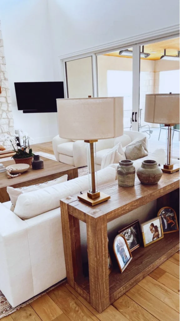 A bright, high-end living room displaying strategic focal point styling. Positioned cleanly behind a cream fabric sofa is a long, rustic wooden console table acting as a structured divider. The table is thoughtfully decorated with a pair of matching contemporary brass-and-glass table lamps, earthy ceramic vases, and framed family photographs on the lower shelf, leading the eye toward an open, bright patio area.