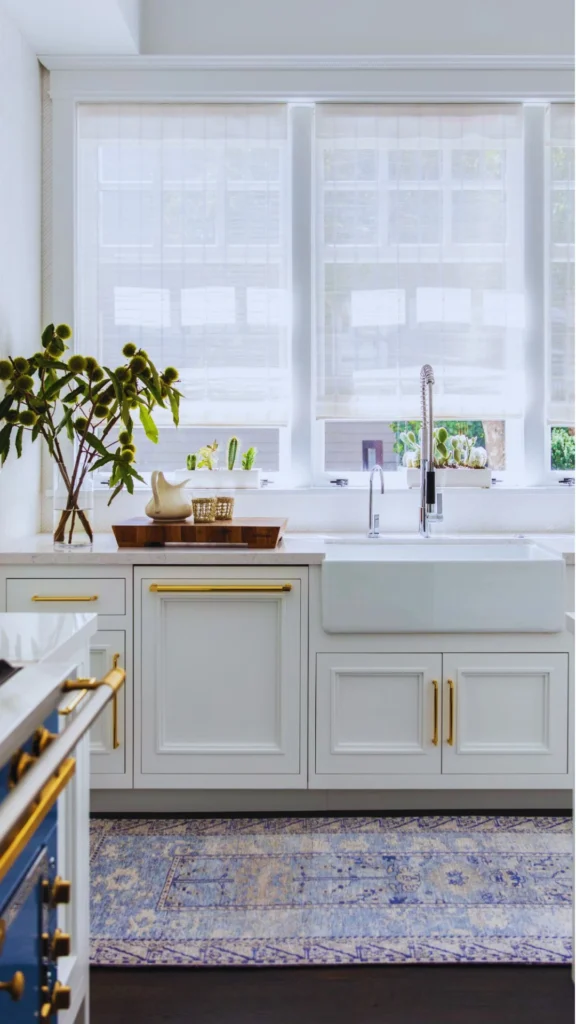 A pristine white luxury kitchen showcasing the precise alignment and strategic orientation of premium gold hardware. Long gold pulls are mounted horizontally along the drawers and integrated dishwasher panel, while identical pulls are mounted vertically on the double doors beneath a white farmhouse apron sink, highlighting clean parallel lines. The balanced space is finished with a chrome commercial-style faucet and a blue vintage area rug.