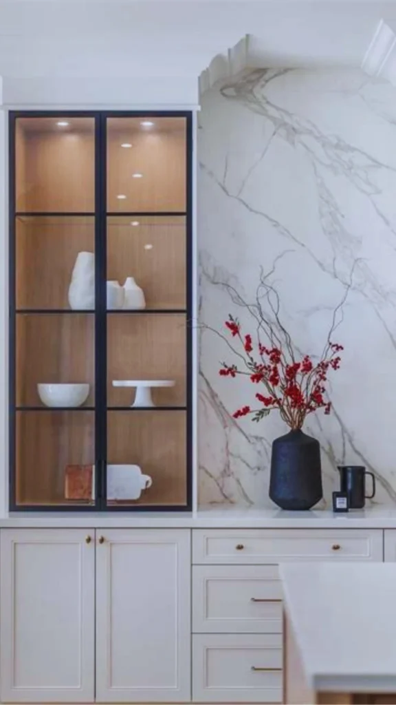 A sophisticated luxury kitchen showing integrated accent lighting inside a glass-front display cabinet. The tall cabinet features a sleek black frame and warm wood interior shelves illuminated by bright overhead puck lights, beautifully showcasing curated white ceramic vases, a bowl, and a cake stand. To the right, a matching white marble backsplash with grey veining frames a dark vase with red berries.