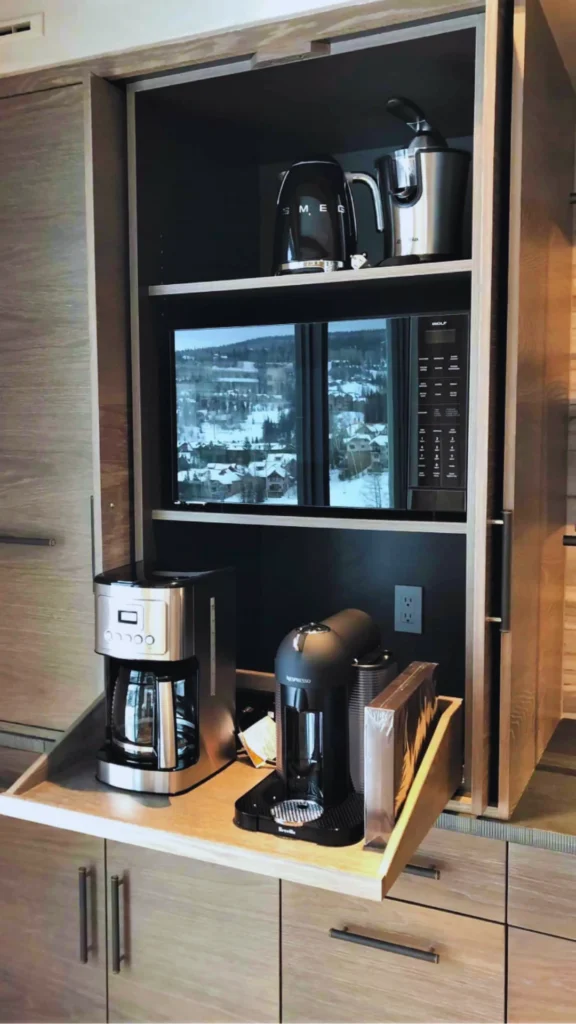 A custom luxury coffee and appliance station built seamlessly into kitchen cabinetry. A heavy wooden pocket door system slides back to reveal an integrated black niche with a pull-out shelf hosting a stainless steel coffee maker and a matte black Nespresso machine plugged into an internal wall socket. The vertical station features an integrated Wolf microwave above, with a Smeg electric kettle and a juicer tucked away neatly on the top shelf.