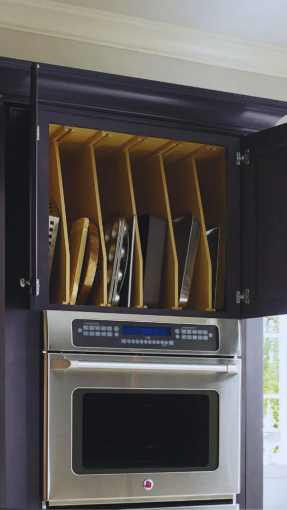 A built-in upper kitchen cabinet displaying an efficient vertical storage solution directly above a stainless steel wall oven. The open dark wood cabinet features custom vertical wooden tray dividers that neatly organize muffin tins, baking sheets, cutting boards, and cookie sheets on their edges rather than stacking them horizontally, keeping the interior looking organized and calm.