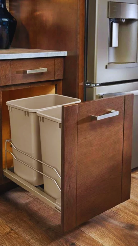 A custom lower kitchen cabinet showing a heavy-duty, integrated pull-out system that prioritizes hygiene and a clutter-free vibe. The warm, stained wood cabinet door slides open on a smooth glider mechanism to reveal a dual bin setup holding two tall, removable beige containers for trash and recycling, neatly tucked away next to a stainless steel refrigerator.