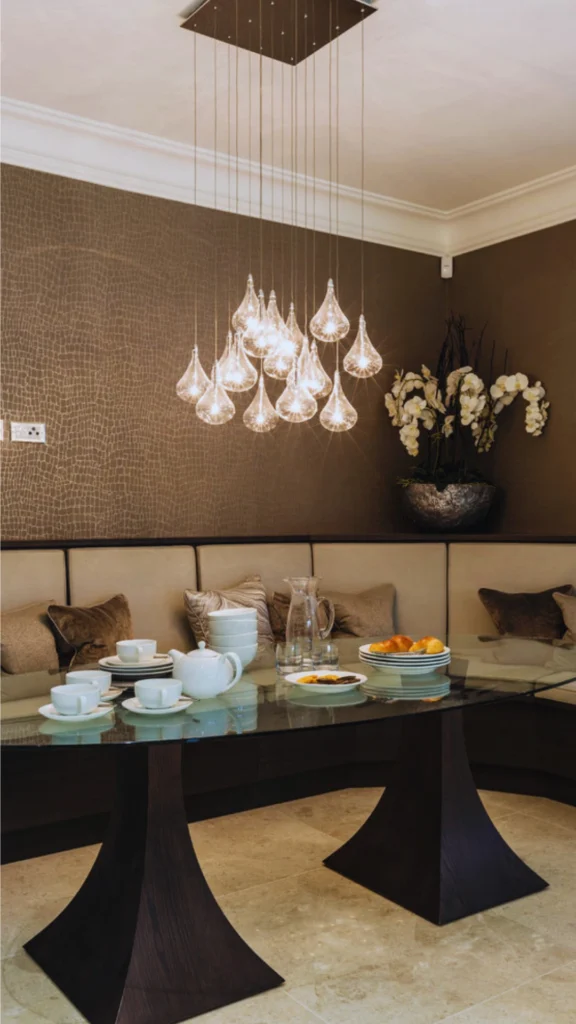 An upscale dining nook demonstrating luxury furniture silhouettes and intentional design. A large, curvilinear glass-top dining table is supported by two sculptural, dark wood geometric bases. The table is surrounded by a custom curved banquette seating area with velvet pillows, all illuminated by a striking modern cluster chandelier featuring cascading glass teardrop pendant lights against textured brown wallpaper.