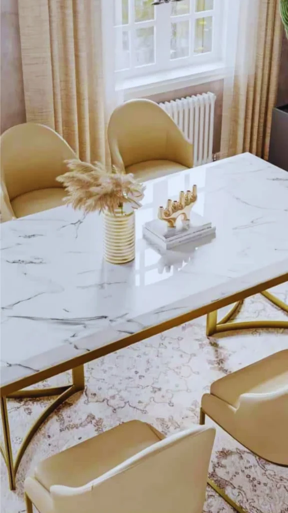 A high-end dining table displaying the cold tactile weight and natural beauty of premium stone. The rectangular table features a polished white marble top with dark, dynamic veining, supported by a structured geometric bright gold metal frame. To showcase the stone's surface, the table is minimally styled with a fluted gold vase holding pampas grass and a sculptural gold object resting on a stack of books, surrounded by cream leather dining chairs.