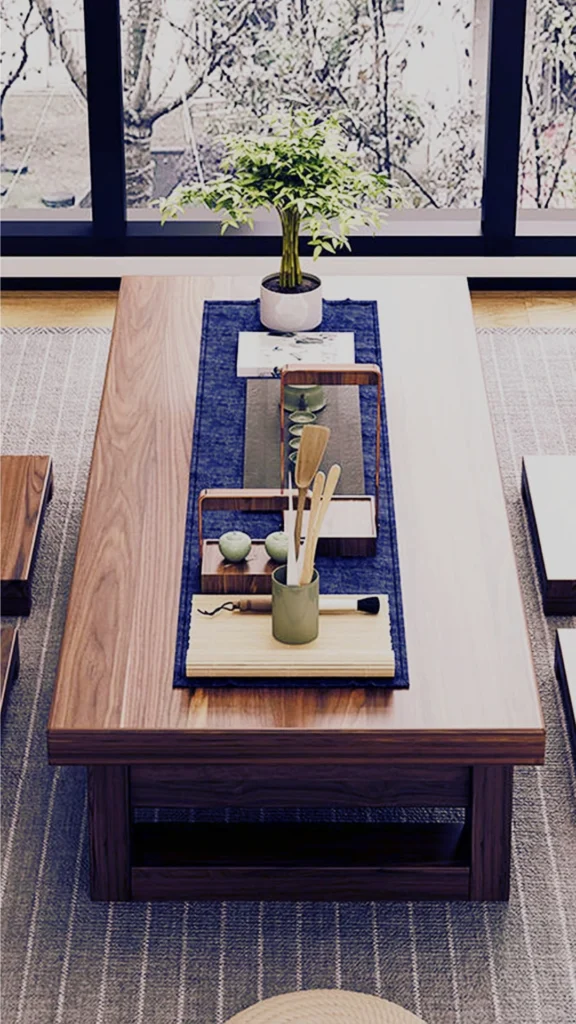 A serene low-profile wooden coffee table embodying a Japanese-inspired low-living aesthetic to make the space feel grounded and intimate. Stretched across the solid wood top is a blue fabric runner styled with a minimalist tea set, bamboo utensils in a container, a small potted green plant, and an art book. The low furniture arrangement sits on a grey textured area rug directly in front of large glass windows looking out to a garden.