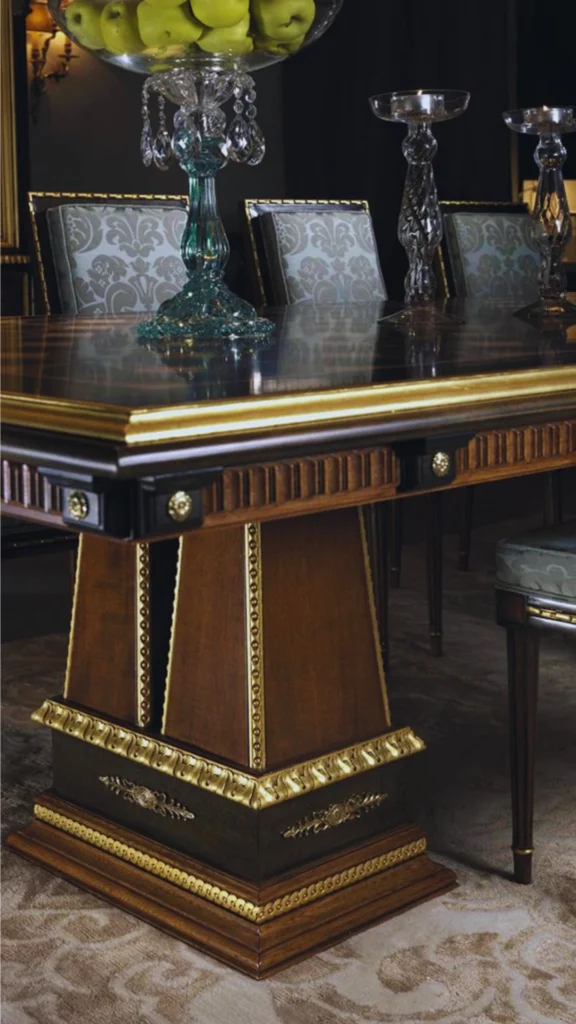 An ornate, traditional luxury dining table illustrating the beauty of historical furniture elements. The heavy, dark wood pedestal base features intricate gilt bronze ormolu mounts and carved gold leaf detailing. On the high-gloss polished wooden tabletop, a crystal centerpiece bowl filled with green apples rests alongside towering glass candleholders, positioned against a backdrop of matching classical upholstered dining chairs.
