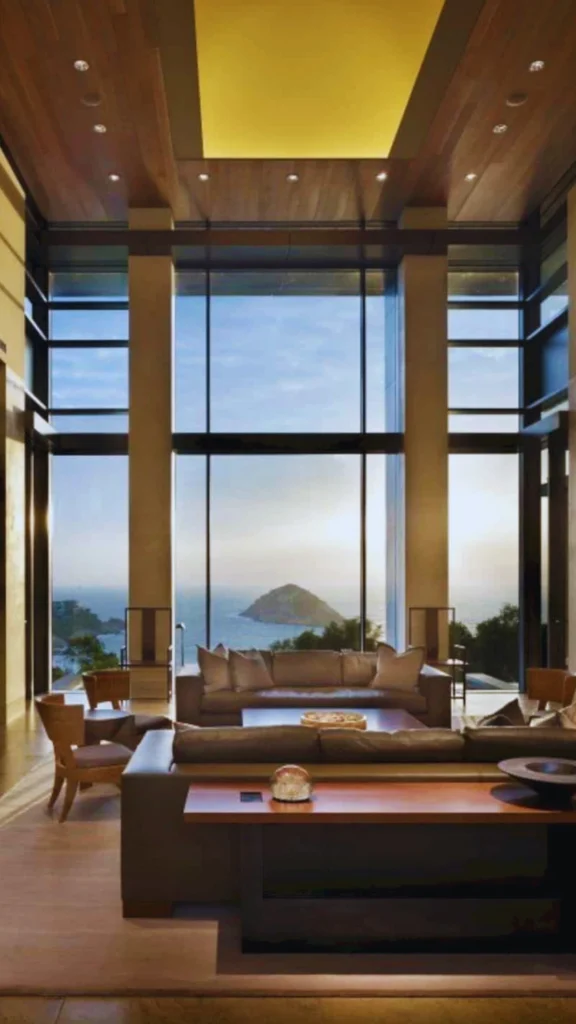 A grand, high-ceilinged luxury living space showcasing the intentional balance of Yin and Yang in Chinese luxury design. Vast floor-to-ceiling windows look out onto a serene ocean bay and a distant island mountain, letting in bright natural light (Yang). Inside, deep, comfortable low-profile brown leather sofas and an anchoring wooden console table (Yin) ground the immense architectural scale, creating a balanced and peaceful atmosphere.