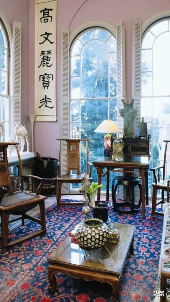 A sunlit, high-end room showcasing classic heritage pieces in a modern Chinese luxury layout. Elegant Ming-style wooden horseshoe armchairs are arranged around a low, glass-top coffee table styled with an ornate ceramic jar. The space features large arched windows, a vibrant blue and red patterned area rug, and a traditional, vertical Chinese calligraphy art scroll hanging prominently on a soft lilac wall.
