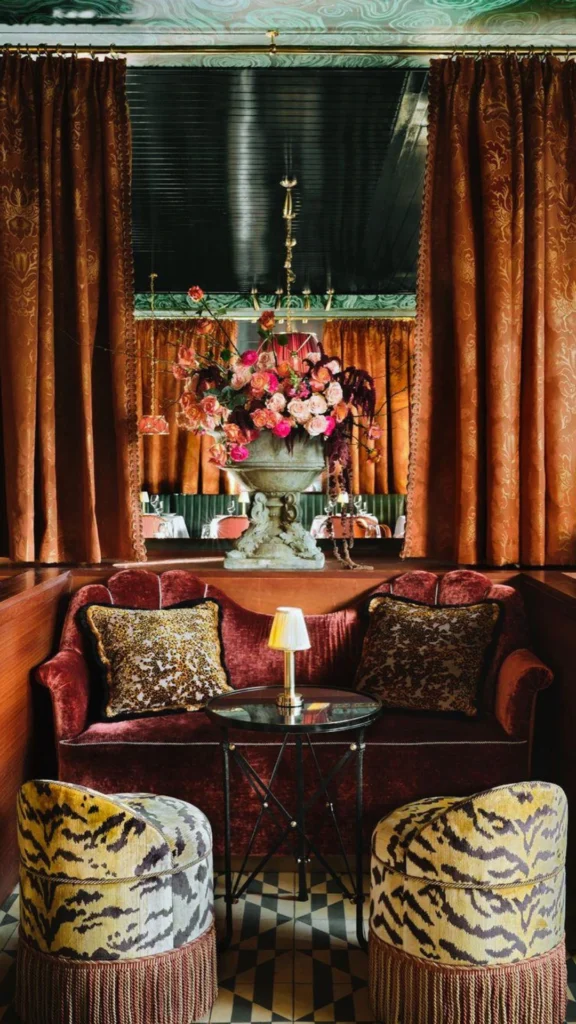 An ultra-luxurious, maximalist interior seating area showing an opulent mix of rich fabrics and textures. A plush velvet tufted sofa in deep burgundy is topped with wild animal print pillows and framed by two round upholstered stools in a striking tiger-stripe pattern with thick fringed trim. In the background, a massive stone urn overflowing with a dramatic floral arrangement of pink and peach roses sits framed by heavy, draped amber brocade curtains.