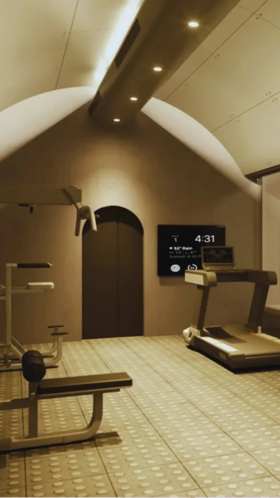 A sleek, high-end home gym inside a luxury underground bunker. Designed for multi-functional wellness, the modern subterranean space features an arched ceiling with integrated recess lighting, a professional treadmill facing an embedded digital screen displaying training data and time, a weight bench station, and durable studded rubber floor tiling in a clean, sandy-neutral tone.