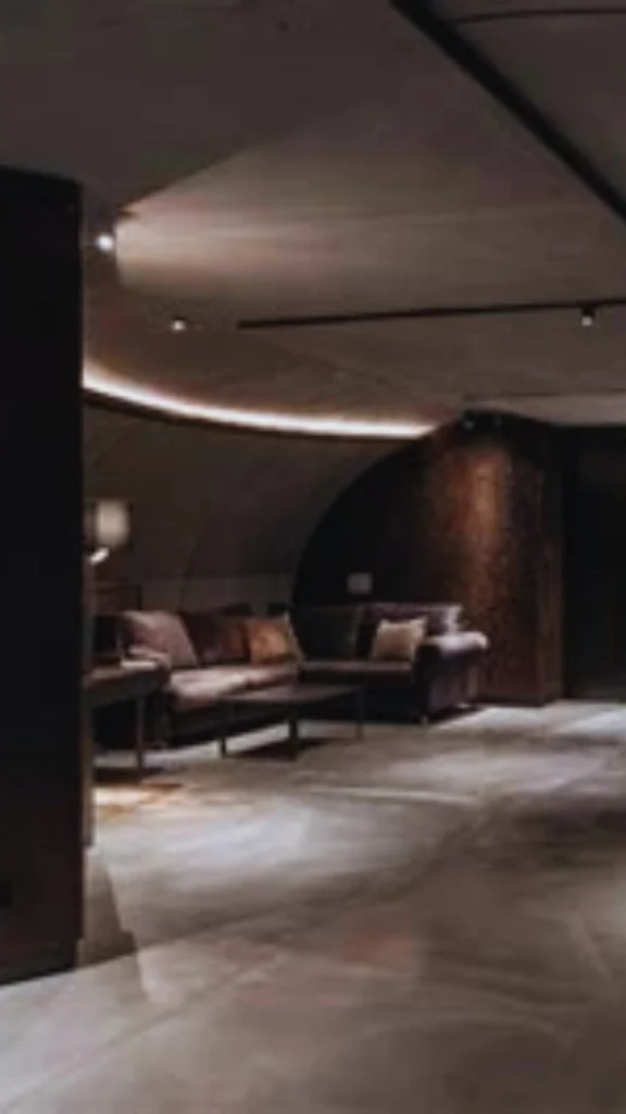 A sophisticated subterranean lounge showcasing the ultimate in luxury climate and comfort control. The space features low-profile leather sectional sofas arranged under a curved concrete ceiling with seamlessly integrated cove lighting and linear HVAC diffusers for invisible air circulation. Polished concrete flooring flows through the open layout, under a perfectly tempered and illuminated atmosphere.