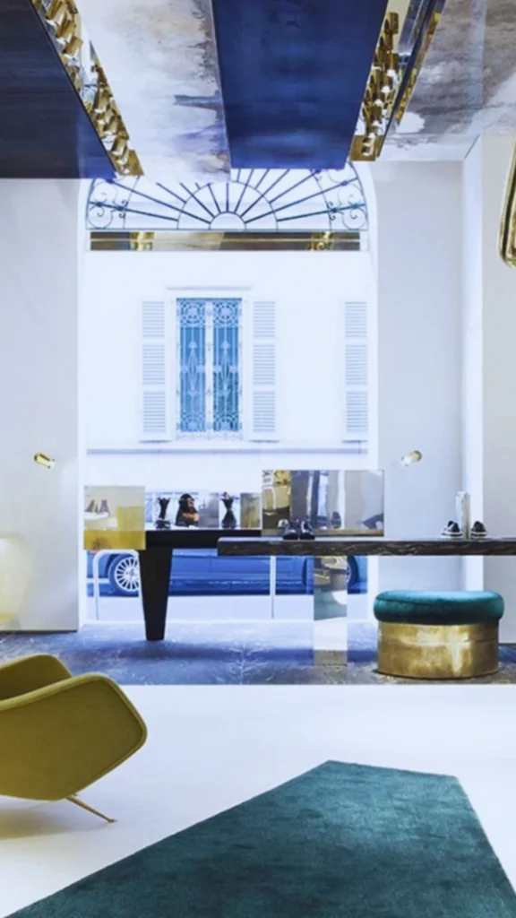 Inside a contemporary high-end luxury boutique looking toward a large street-facing window. A modern dark slab display table stands before the window, presenting designer footwear. The interior layout features premium textures including a teal velvet and brass ottoman, a mustard-yellow accent chair, a deep green geometric rug on light floors, and a dramatic ceiling accented with polished dark and golden metallic panels.