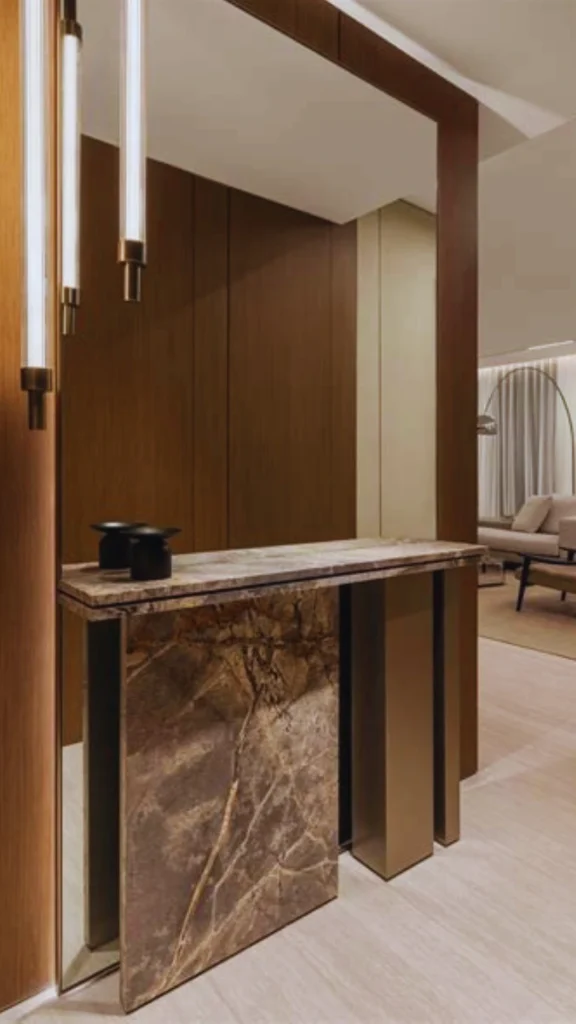 An elegant high-end entryway showcasing dense, premium textures that communicate quality. A heavy console table crafted from dark brown marble with dramatic natural veining stands against a warm wood-paneled wall. Sleek, vertical brass light fixtures illuminate the substantial stone table, which rests on a light oak floor, perfectly demonstrating the impact of authentic materials on price perception.