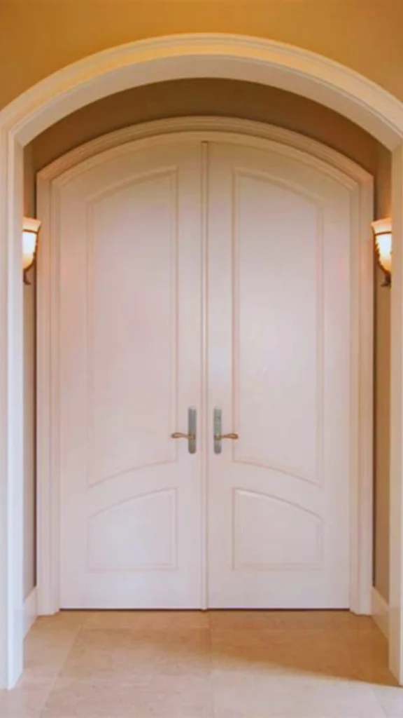 An artful interior entrance featuring a pair of elegant white double doors housed inside a beautiful arched architectural frame. The custom doors feature deep panel molding with sweeping curved lines along the bottom panels. Accenting the center are two substantial, vertical silver-toned lever handles with oversized backplates that act as the door's jewelry, illuminated by warm, symmetrical wall sconces on either side of the hallway.