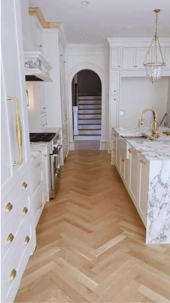 A luxury custom kitchen showcasing a sophisticated chevron or herringbone wood floor pattern that channels a classic French chateau vibe. The light oak planks create a dynamic geometric runway down the center of the kitchen, pulling the eye forward toward an elegant white arched doorway. The statement flooring serves as a beautiful warm focal point, beautifully balancing the surrounding white cabinetry and dramatic marble countertops.