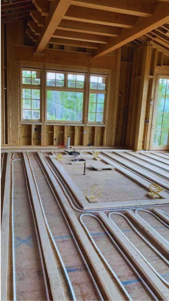 An inside look at the subfloor installation of a whole-house hydronic radiant floor heating system before the luxury flooring is laid. Sinuous white PEX tubing loops systematically through precise wooden channels cut into the subfloor panels. The structural groundwork is set inside a high-end home under construction, featuring exposed timber wall framing, dramatic raw wood ceiling beams, and a triple-window array.