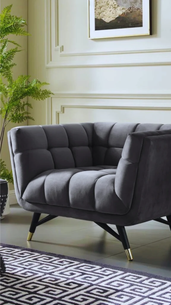 A sophisticated living room composition highlighting a sculptural luxury armchair. The velvet barrel-curved armchair features a deep charcoal grey hue, deep channels, and a tufted geometric pattern. Angled black wooden legs with striking polished gold metal tips provide an artistic base. The chair is positioned as a focal point against a cream wall with classic picture frame molding and a monochromatic geometric rug in the foreground.