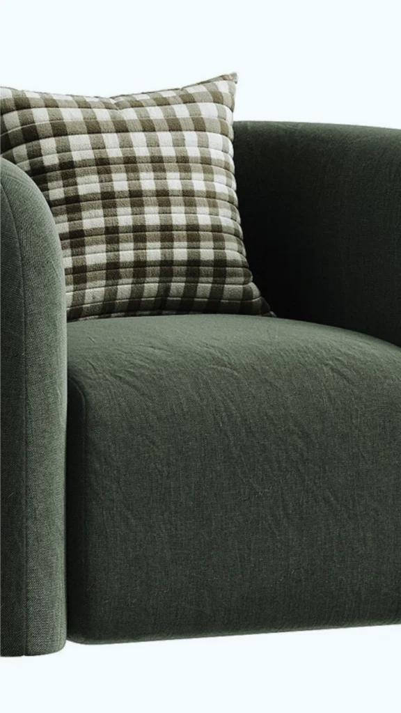 A close-up shot showcasing the "soft luxury" aesthetic with a plush, rounded armchair designed to soften the atmosphere. The chair features an enveloping, curved armrest upholstered in a textured forest green linen fabric. Nestled inside the deep seat is a square accent pillow with a classic brown and cream gingham plaid pattern, emphasizing physical comfort and human-centric design.