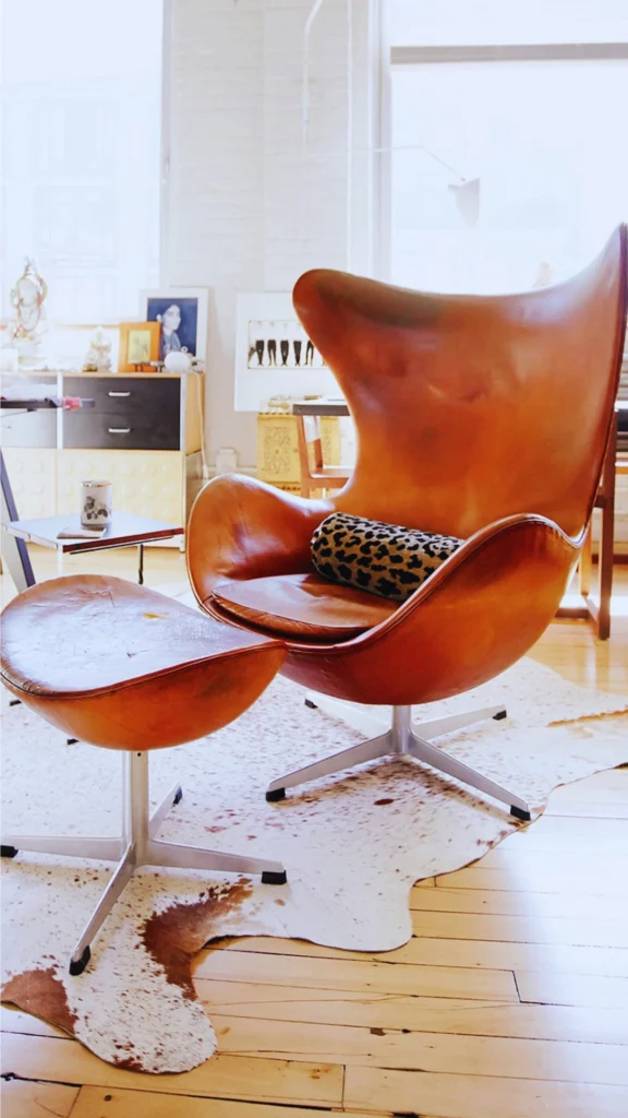 An iconic piece of design history anchoring a sophisticated room with its quiet authority. The mid-century modern Egg Chair and matching footstool, designed by Arne Jacobsen, are beautifully upholstered in a rich, patinated cognac tan leather. Resting on metallic four-star swivel bases, the heritage lounge set features a contrasting leopard-print bolster pillow and sits on a brown and white cowhide rug atop light hardwood floors.