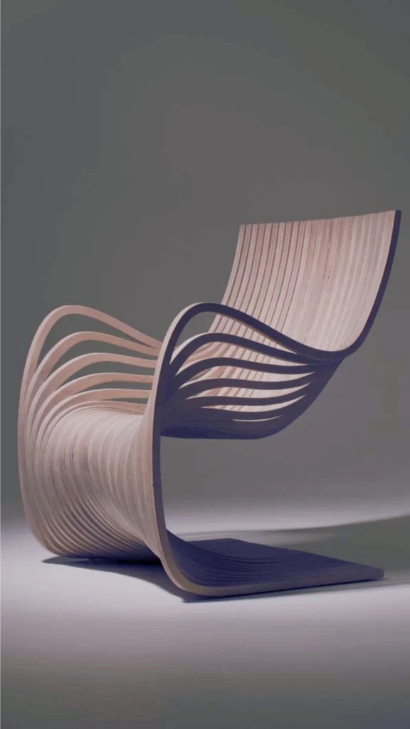 An avant-garde luxury accent chair exemplifying how tactile materiality can create a powerful textural contrast in high-end spaces. The chair is crafted out of thin, bent plywood strips that fan out to form a fluid, undulating wave shape. This striking sculptural configuration provides deep contours for the seat, backrest, and dramatic looping armrests, resting on a cantilevered wooden base against a minimalist grey backdrop.