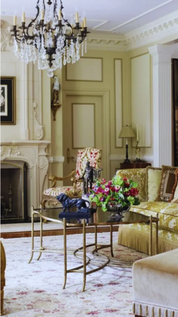 A traditional luxury living room rich in architectural detailing that provides a sense of structure and permanence. The pale yellow-green walls are adorned with classic framed wall divisions and subtle moldings, complemented by intricate white ceiling cornices and a fluted pillar column. An ornate carved stone fireplace mantel and an elegant crystal chandelier ground the seating area, which is filled with patterned damask furniture and an antique gold-framed glass coffee table.