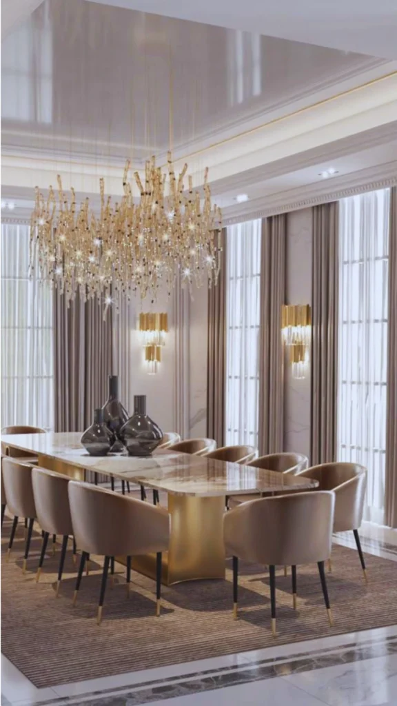A grand neo-classical dining room showcasing a layered luxury lighting scheme anchored by a massive, sculptural gold crystal chandelier that resembles shimmering, organic branches hanging from a glossy recessed ceiling coffer. The ambient glow is balanced by architectural downlights and twin tiered gold wall sconces, illuminating an upscale marble dining table with modern velvet chairs against floor-to-ceiling windows.