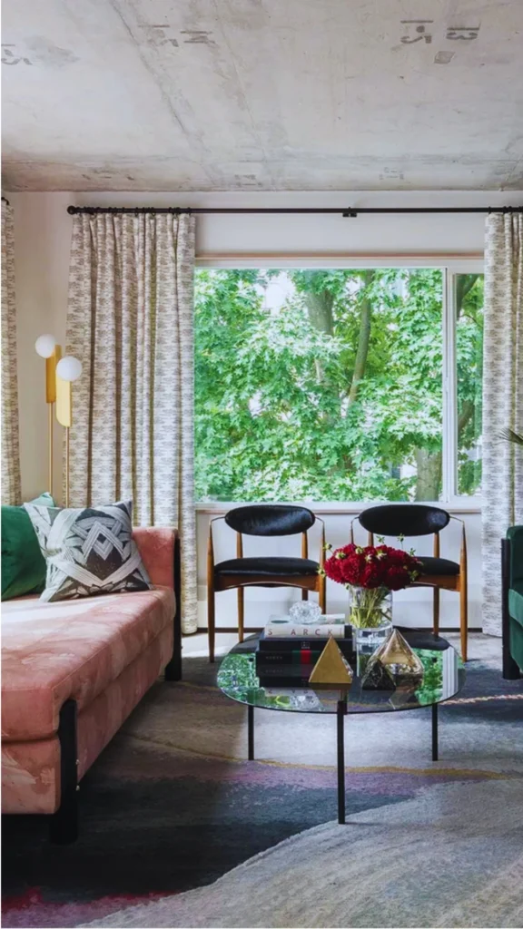 An edited, quietly luxurious contemporary classic living room composition framing a large window with vibrant green trees outside. A balanced pair of mid-century wooden armchairs with black upholstered seats rests symmetrically below the windowsill. In the center, a round glass coffee table is intentionally styled with a stacked hardback book, a brass pyramid sculpture, and a vase of deep red flowers, pulling together the rich velvet textures of a dusty pink sofa and a multicolored abstract area rug.