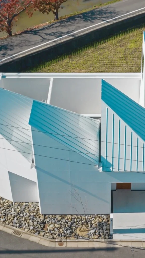 An aerial view of an ultra-modern luxury home showcasing a sculptural roof masterpiece designed to serve as a signature architectural statement. The property features a striking, multi-angled roof structure finished in vibrant teal blue-green metal paneling that slopes dramatically across a stark white concrete facade. The unconventional geometric design cuts sharp architectural lines against the surrounding landscape, capturing dynamic light and shadow.