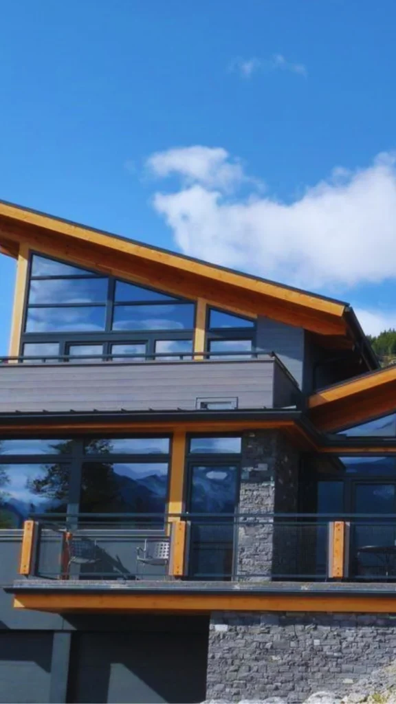 A luxury multi-level modern mountain home showcasing a striking architectural profile that perfectly complements open floor plans. The structure features an upward-sloping, shed-style roofline framed in warm natural wood trim, creating an asymmetric silhouette against a bright blue sky. Large, glass-windowed clerestory gables reflect the surrounding landscape, while premium glass-enclosed outdoor entertainment decks extend the functional living space.