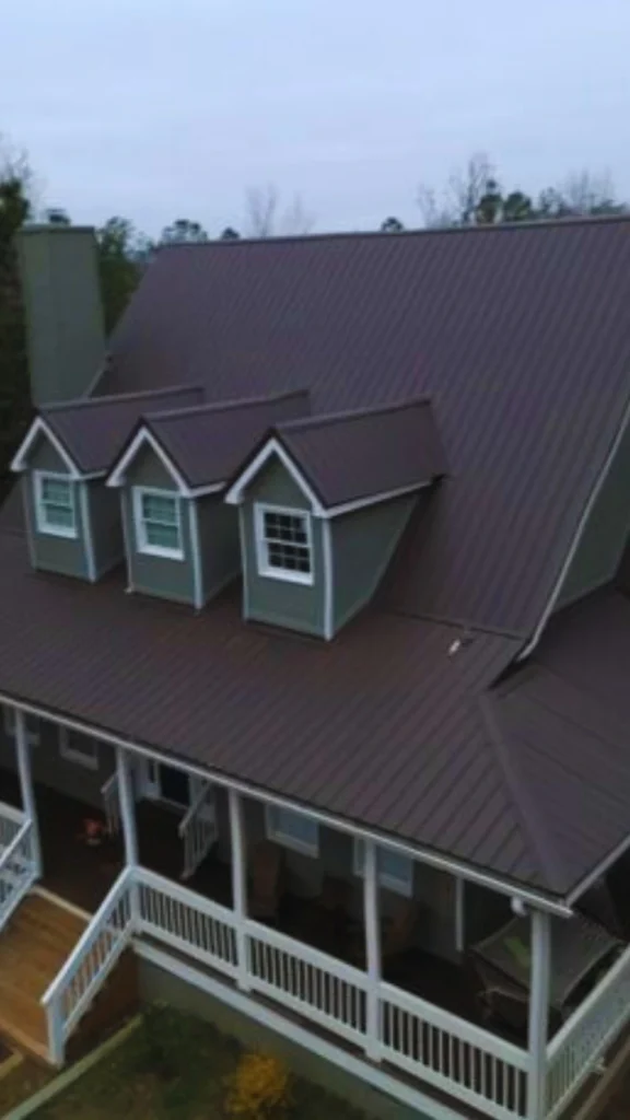 A high-angle aerial view of a stately residential property showcasing a grand, steeply pitched roof designed to crown the home with traditional charm. The expansive roof is finished in clean vertical panels of a deep, warm burnt sienna or dark bronze hue, which beautifully echoes the prestige of classic estate roofing. Three symmetrical, small gabled dormer windows with crisp white trim break up the upper roofline, adding visual rhythm above a wide, wrap-around porch with white railings.