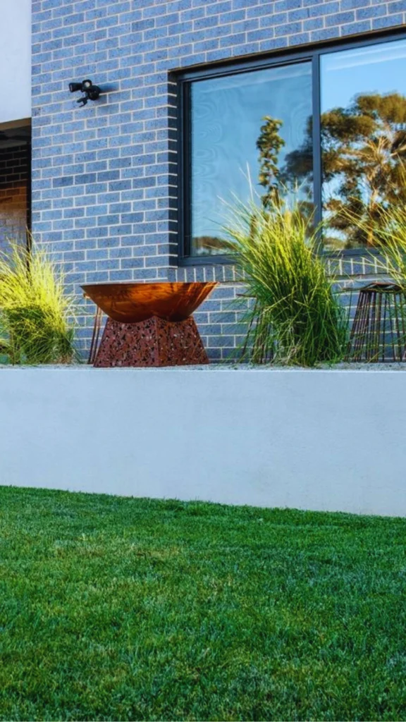 A professionally designed modern residential landscape showing a seamless integration of architecture and curated greenery. Set against a textured blue-grey brick facade with a large black-framed window, a custom-built elevated concrete planter bed holds clusters of vibrant, tall ornamental grasses. Centered in the design is a sculptural, rusted Corten steel fire bowl or planter resting on an intricate, geometric pattern base, demonstrating an effortless and intentional sanctuary aesthetic.