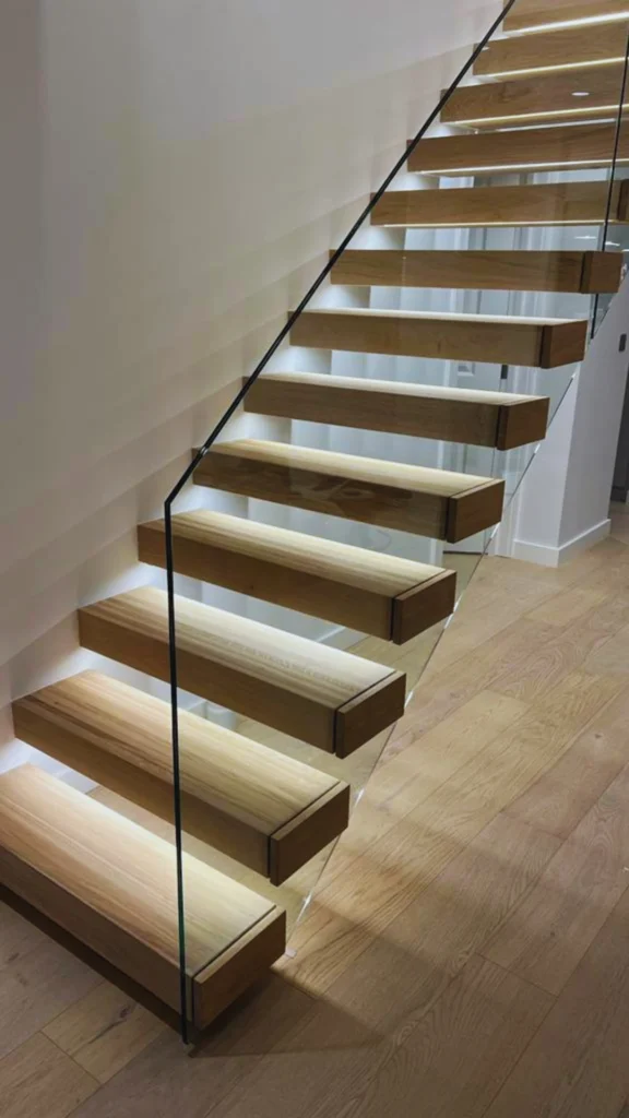 A premium modern floating staircase demonstrating how a frameless glass balustrade allows an interior design to breathe. The structure features thick, open-tread light oak wood steps cantilevered from a minimalist white wall. With no visible posts or rails interrupting the sightline, the seamless glass panel runs smoothly alongside the flight, while integrated LED lighting hidden under each tread casts a warm, soft glow that maximizes light flow through the space.