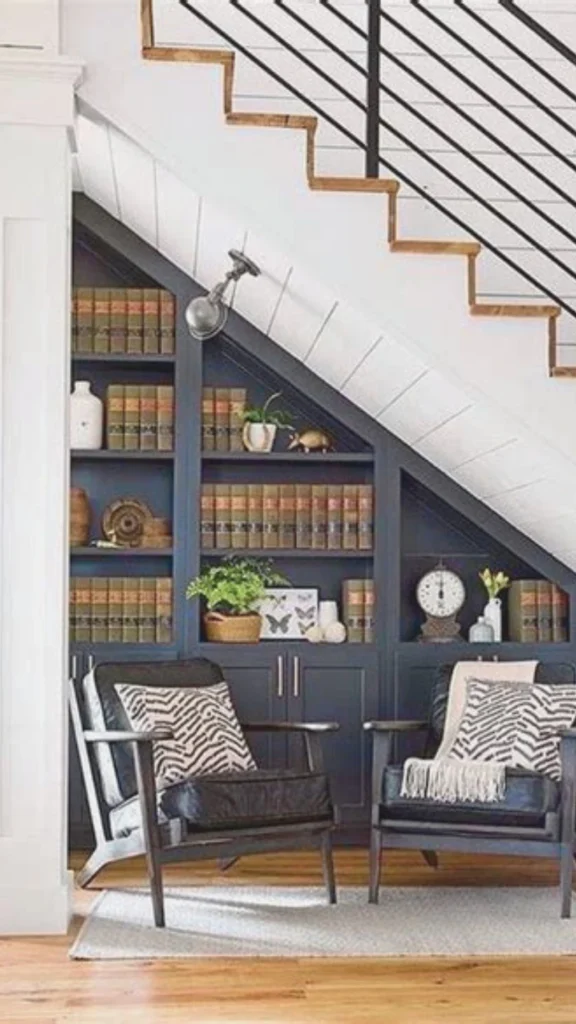 A premium residential staircase design demonstrating a completely reimagined under-stair space utilized for both form and function. Built directly into the slope beneath the white wood paneled stairs is a custom, dark charcoal grey library bookcase filled with matching hardbound books, small potted plants, and decor accents. Two modern black wooden armchairs with patterned throw pillows face the built-in shelving on a light area rug, creating a sophisticated and cozy reading nook that maximizes every square metre.