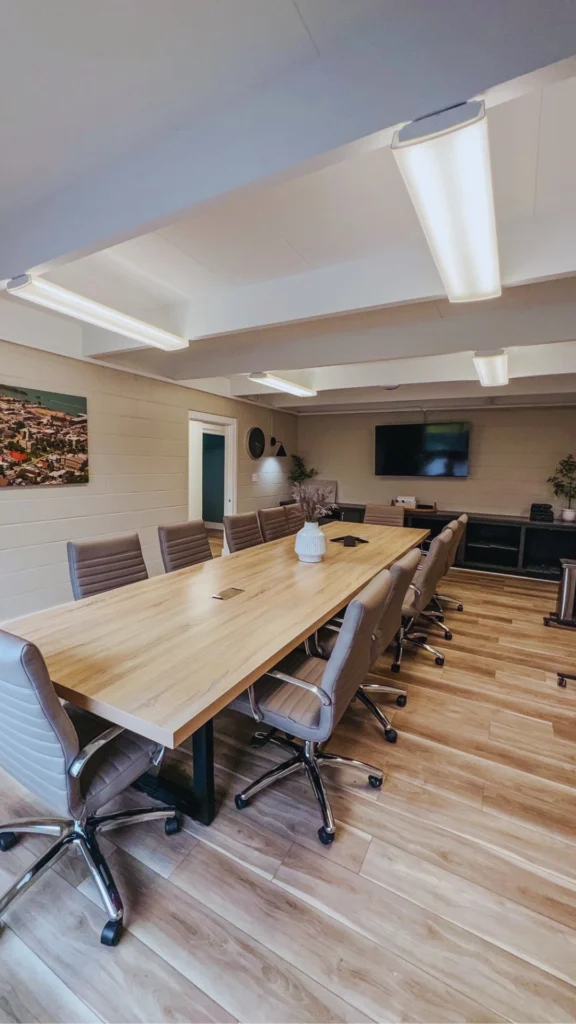 Interior view of a contemporary professional meeting room or boardroom setup. A large, rectangular light wood table stretches through the room, lined with ten matching high-back beige rolling office chairs. The space is finished with light-colored faux brick walls, a wooden plank floor, and a large flat-screen TV mounted on the far wall above low-profile dark storage cabinetry.