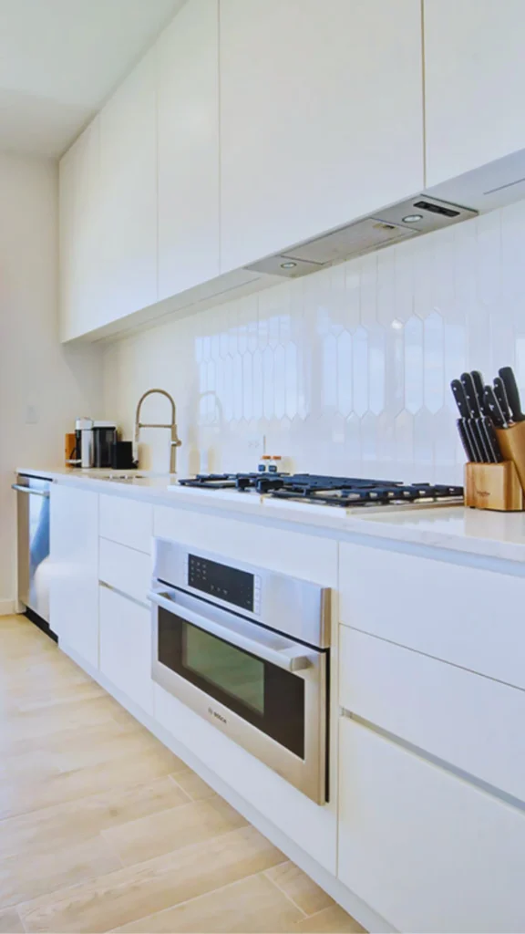A side-angle view of a contemporary, minimalist white galley kitchen. The setup features long, handleless white upper and lower cabinets, light wood-look floor tiles, a stainless steel oven, a gas stovetop, and a white tiled backsplash with a subtle vertical geometric pattern. A wooden block knife set sits on the clean white quartz countertop in the foreground.