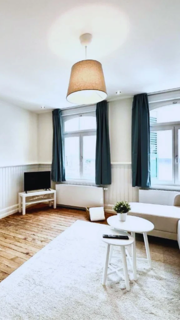 Interior view of a bright, modern minimalist apartment living room with character features. The space displays natural wood plank flooring, light-colored walls, and two large windows with floor-length slate blue curtains. A soft cream area rug sits under a pair of round white wooden nesting tables and the edge of a light fabric sofa, while a compact television sits on a low table in the corner under a large hanging pendant lamp.